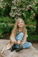 gelukkig meisje kind met lang golvend bruin haar- zit Aan een steen in voorjaar in een bloeiend park en glimlacht foto