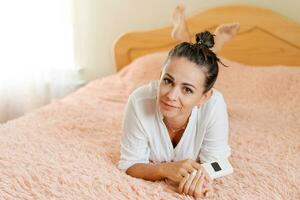 schattig vrouw aan het liegen Aan de bed met een telefoon in haar hand- Aan een zacht roze deken foto