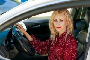 mooi geslaagd blond vrouw in een rood jurk zittend in een auto achter de wiel foto