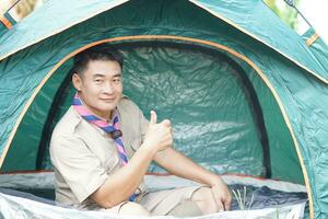knap Aziatisch Mens draagt jongen verkenner uniform zit in camping tent. concept, accommodatie voor tijdelijk buitenshuis slapen net zo verkenner wandelen werkzaamheid. verkenner Gaan camping. foto