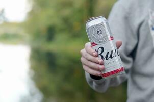 kan van budweiser knop bier in mannetje hand- Aan een rivier- en groen bomen wazig achtergrond foto