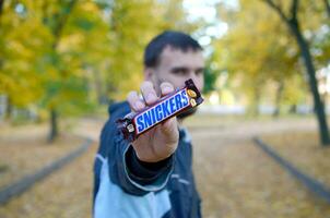 Charkov, Oekraïne - oktober 8, 2019 een jong Kaukasisch gebaard Mens shows grinnikt chocola bar in bruin omhulsel in herfst park. grinnikt chocola vervaardigd door Mars foto