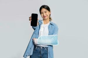 Aziatisch vrouw geduldig met gebroken arm na ongeluk en vervelend arm gips voor behandeling. ze shows wit scherm smartphone geïsoleerd Aan wit achtergrond. bespotten omhoog. foto