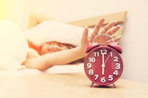 jonge vrouw fronsend in bed en hand op rode wekker. foto