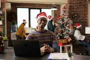 glimlachen Afrikaanse Amerikaans bedrijf werknemer vervelend de kerstman hoed en winter gebreid trui zittend Bij kantoor bureau portret. vrolijk Mens zakelijke arbeider in versierd Kerstmis werkplaats op zoek Bij camera foto