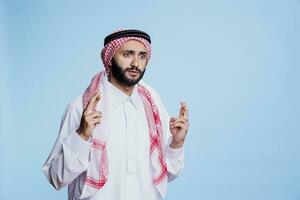 bang Mens gekleed in cultureel moslim kleren staand met gekruiste vingers. doodsbang Arabisch persoon met ogen breed Open poseren met bijgelovig gebaar voor geluk in studio Aan blauw achtergrond foto