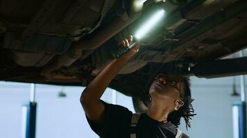 licentie monteur staand onder geschorst auto in garage werkruimte, gebruik makend van werk licht naar controleren voor schade en tablet naar bestellen nieuw vervangen onderdelen na vinden problemen gedurende jaar- onderhoud foto