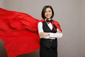 glimlachen zelfverzekerd Aziatisch receptioniste staand in fladderend supervrouw rood kaap en op zoek Bij camera. vrolijk serveerster met armen gekruiste poseren in superheld mantel portret foto