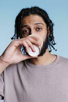 Arabisch Mens drinken koffie naar Gaan en op zoek Bij camera met kalmte gelaats uitdrukking portret. jong persoon Holding nemen uit cappuccino papier kop terwijl poseren Aan blauw achtergrond foto