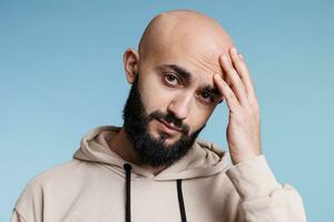 Arabisch Mens aanraken hoofd in pijn terwijl hebben migraine en op zoek Bij camera. jong persoon lijden van hoofdpijn terwijl Holding hand- Aan voorhoofd en poseren voor studio portret foto