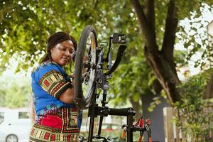 gefocust gezond Afrikaanse Amerikaans vrouw herstellen haar eigen fiets in werf en het uitvoeren van jaar- buitenshuis onderhoud gebruik makend van deskundige hulpmiddelen. vrouw fietser inspecteert en reparaties fiets pedalen. foto