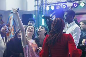 vrolijk clubbezoekers nemen groep foto Aan smartphone terwijl feesten Aan druk nachtclub dansvloer met lichten. zorgeloos glimlachen vrouw maken mobiel telefoon selfie met vrienden in club