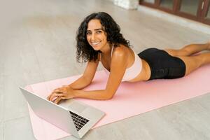 glimlachend brunette vrouw met gekruld haren chillen na online opleiding Aan yoga mat. poseren Aan tropisch terras. foto