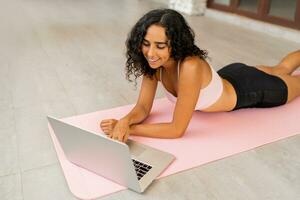 glimlachend brunette vrouw met gekruld haren chillen na online opleiding Aan yoga mat. poseren Aan tropisch terras. foto