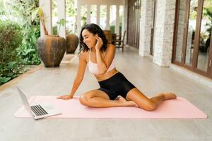 bevallig vrouw in elegant sport kleding op zoek online opleiding en zittend Aan yoga mat Aan terras. lichaamsverzorging en welness concept. foto