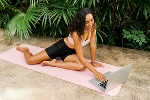 bevallig vrouw in elegant sport kleding op zoek online opleiding en zittend Aan yoga mat over- tropisch tuin. lichaamsverzorging en welness concept. foto