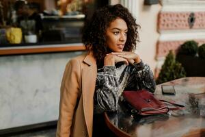 buitenshuis portret van mooi glimlachen zwart vrouw met elegant afro haren zittend in cafe in Parijs. vervelend modieus fluweel jurk en beige jas. luxe tas. foto