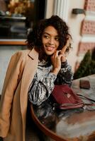 buitenshuis portret van mooi glimlachen zwart vrouw met elegant afro haren zittend in cafe in Parijs. vervelend modieus fluweel jurk en beige jas. luxe tas. foto