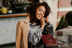 buitenshuis portret van mooi glimlachen zwart vrouw met elegant afro haren zittend in cafe in Parijs. vervelend modieus fluweel jurk en beige jas. luxe tas. foto