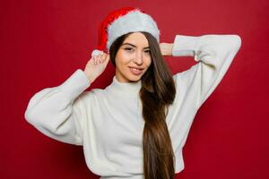 gelukkig vrouw met verbazingwekkend lang haren in de kerstman hoed poseren in studio Aan rood achtergrond. foto