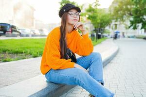 buitenshuis levensstijl beeld van romantisch dromerig vrouw zittend Aan trottoir en genieten van avond . vervelend elegant jeans en oranje trui. foto