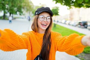 blij glimlachen brunette meisje in ronde bril en modieus oranje gebreid trui maken zelf portret door camera. zonnig avond . foto