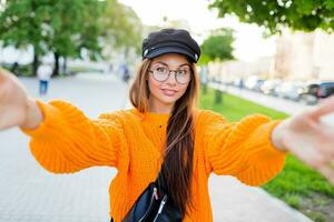 blij glimlachen brunette meisje in ronde bril en modieus oranje gebreid trui maken zelf portret door camera. zonnig avond . foto