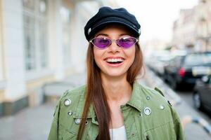 speels vrouw poseren buitenshuis . vervelend modieus Purper retro bril , zwart pet . dichtbij omhoog portret . winderig haren. levensstijl afbeelding. foto