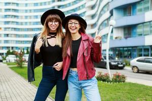 twee zorgeloos glimlachen Dames poseren over- modern stad achtergrond. vervelend wol hoed , leer jasje en jeans. vrienden knuffelen. gelukkig emoties . foto