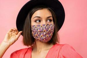 dichtbij omhoog portret van mooi vrouw gekleed beschermend elegant gezicht masker. vervelend zwart hoed en zonnebril. poseren over- roze achtergrond in studio. foto