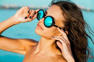 dichtbij omhoog portret van elegant mooi sexy meisje in bril en met nat haar- Aan een zonnig strand met blauw water. zonnebaden en genieten de rust uit. foto