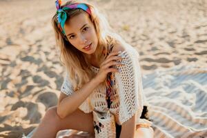 warm zomer portret van verbijsterend bevallig vrouw genieten van zonsondergang in de buurt oceaan, zittend Aan zand. perfect glimlach, vervelend wit boho jurk en kleurrijk hoofdband. foto