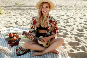 romantisch blond vrouw in rietje hoed zittend Aan Hoes Aan de strand en spelen ukulele gitaar. foto