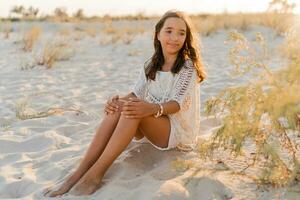 zomer foto van klein meisje in elegant boho kleding poseren Aan de strand. warm zonsondergang kleuren. wacht en reizen concept.