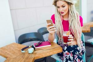 modieus vrouw met dreadlocks gebruik makend van slim telefoon, berichten foto's in stad cafe. foto