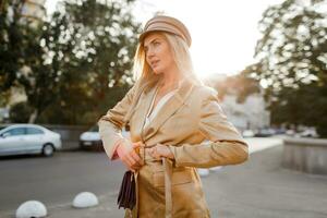 elegant blond vrouw in beige pet en jasje wandelen Aan de straat. herfst Look. zonsondergang lift. elegant tas. foto