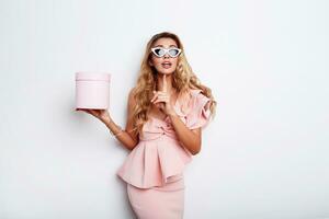verleidelijk blond vrouw Holding geschenk doos en poseren in roze jurk over- wit achtergrond . boodschappen doen en vieren concept. modieus zonnebril. studio schot. foto