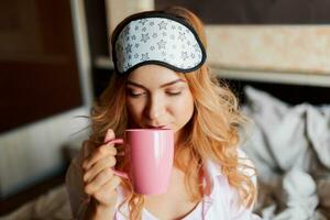 mooi vrouw met openhartig glimlach poseren in oog masker Bij huis en drinken heet thee. knus huis atmosfeer. foto