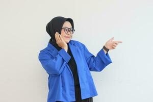 mooi jong Aziatisch moslim vrouw in bril, hijab en vervelend blauw blazer glimlachen zelfverzekerd en gelukkig met beide handen richten zijwaarts foto