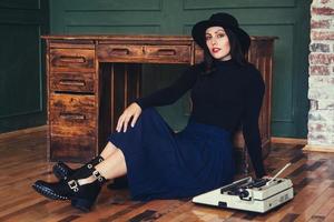 mooie vrouw in een hoed zit in de buurt van eiken tafel met vintage typemachine foto