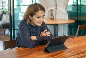 Aziatisch vrouw zittend houten tafel gebruik makend van digitaal tablet wacht onine films stroom of boodschappen doen online Bij leven kamer, gelukkig genieten van met vermaak media online Aan weekenden. foto