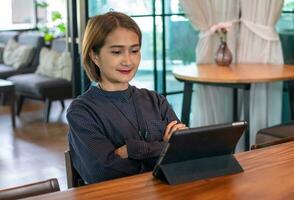 Aziatisch vrouw zittend houten tafel gebruik makend van digitaal tablet wacht onine films stroom of boodschappen doen online Bij leven kamer, gelukkig genieten van met vermaak media online Aan weekenden. foto