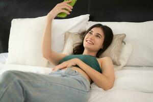 gelukkig jong Aziatisch vrouw gebruik makend van smartphone aan het liegen Aan bed in de slaapkamer van huis. foto