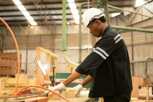 jong Mens arbeider werk in een houtbewerking fabriek, werken met hout zagen en snijdend machines naar produceren houten lakens voor maken pallets foto