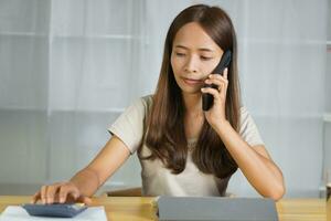 Aziatisch vrouw werken Bij huis gebruik uw mobiel telefoon naar contact de team. foto