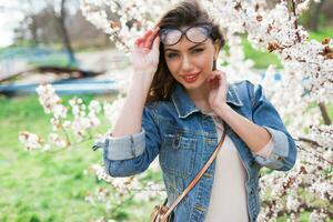 sexy vrouw in jeans jasje, rood vol lippen, golvend kapsel poseren in de buurt bloem boom in voorjaar dag. foto