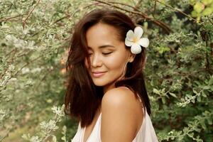 bevallig Aziatisch vrouw met perfect huid en hibiscus bloem in haren poseren over- hout muur en tropisch planten. zomer mode portret. foto