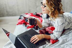 elegant slim vrolijk vrouw in bril gebruik makend van laptop , aan het liegen Aan knus bed, drinken koffie. foto