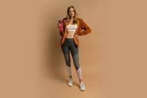 mooi blond vrouw in sportief kleding poseren Aan beige achtergrond. Holding fles van water. vol lengte. foto