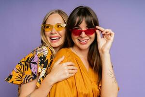 dichtbij omhoog portret van twee elegant Dames in zonnebril en modieus zomer kleren poseren Aan Purper bakgrond in studio. foto